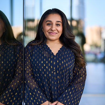 A photo of Shani Dhanda smiling. She has long, dark hair and wears a blue top with gold pattern.