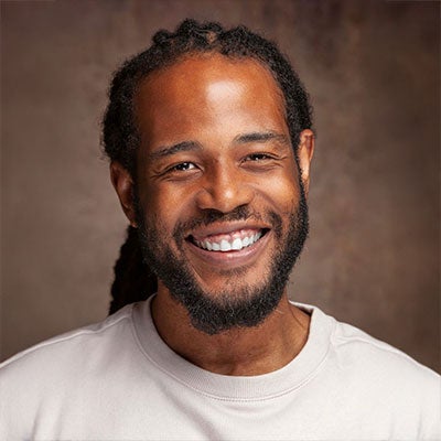 A photo of Ekow Otoo smiling. His dreadlocks are tied back and he has a beard. He is wearing a white t-shirt.
