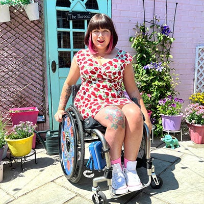 A photo of Nina Tame smiling. She has medium length brown hair with pink streaks. She wears a white dress with a red floral pattern. 