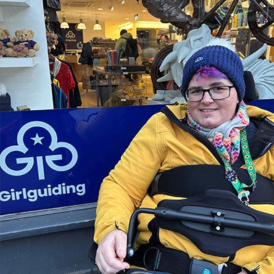 A photo of Zahari Brimacombe smiling in front of a Girlguiding sign. They are wearing a blue beanie, black-rimmed glasses and a yellow jacket.