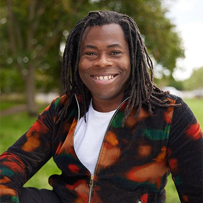 A photo of Ade Adepitan smiling. He has brown dreadlocks and is wearing a black and orange jumper.