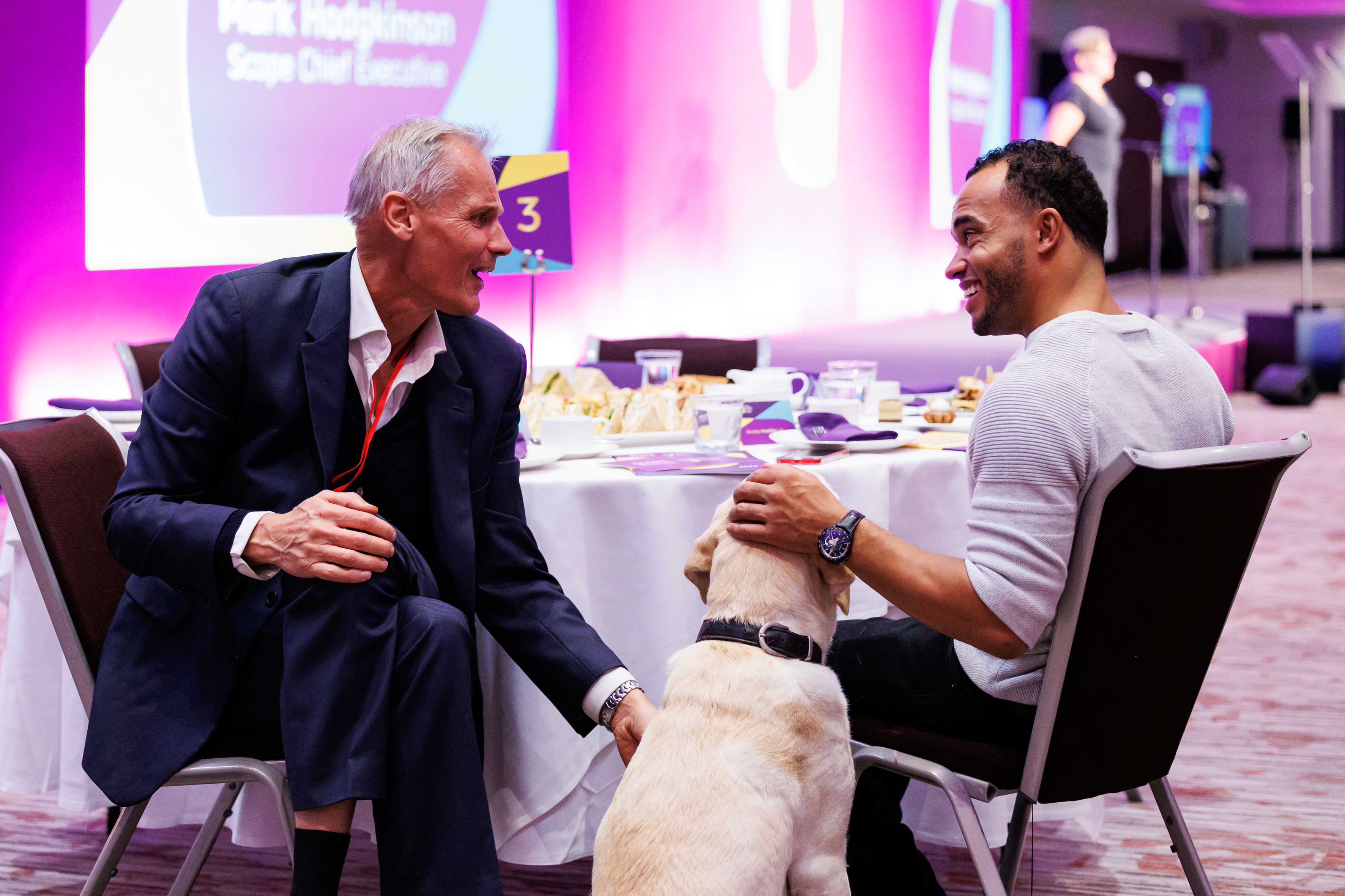 2 people sitting at a table stroking a guide dog