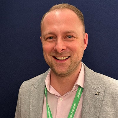 A photo of Andy Cotter smiling. He has short, light-brown hair, and wears a pink shirt and grey blazer. 