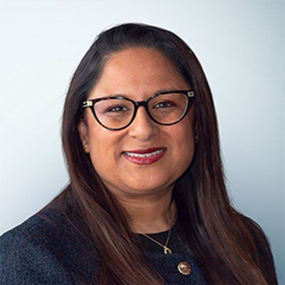 A photo of Reena Parmar smiling. She has long, dark hair and wears black-rimmed glasses and a black jacket. 