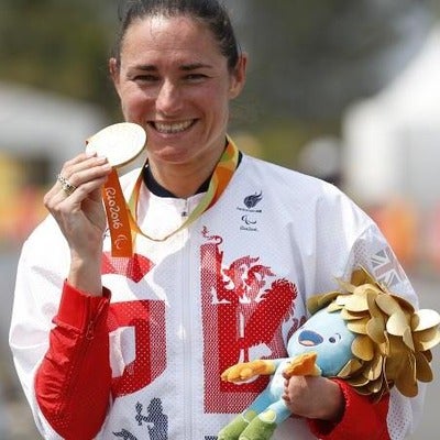 A photo of Sarah Storey smiling and holding up her gold medal. Her hair is tied back and she wears a white and red Paralympic kit.