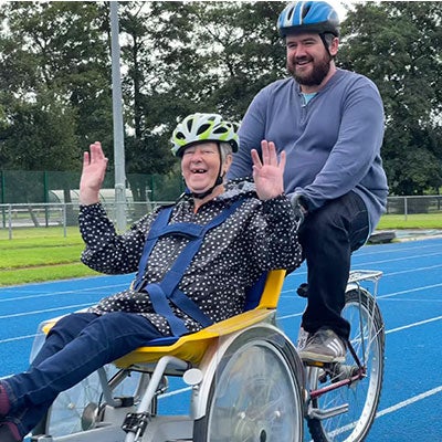 A man riding a bicycle with a woman in a wheelchair, both smiling at the camera. 